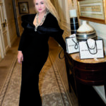 Portrait en pied d'Anna K, fondatrice de la marque "Boutique Eco Diamant", dans le couloir d'un établissement de standing. Elle porte une robe de soirée longue et noire à grand nœud de satin et un collier serti. Des sacs de shopping de sa marque sont posés sur la console en bois à ses côtés.