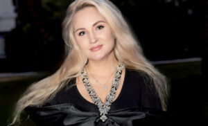 Portrait à mi-corps d'Anna K, fondatrice de la marque "Boutique Eco Diamant", posant en extérieur en soirée. Elle porte une robe noire ajustée ornée d'un grand nœud en satin au décolleté, un collier plastron étincelant et un bracelet rivière. Elle tient à la main des sacs de shopping blancs au nom de sa marque "Boutique Eco Diamant", appuyée près de l'aile d'une voiture de collection.