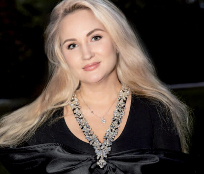 Portrait à mi-corps d'Anna K, fondatrice de la marque "Boutique Eco Diamant", posant en extérieur en soirée. Elle porte une robe noire ajustée ornée d'un grand nœud en satin au décolleté, un collier plastron étincelant et un bracelet rivière. Elle tient à la main des sacs de shopping blancs au nom de sa marque "Boutique Eco Diamant", appuyée près de l'aile d'une voiture de collection.