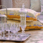 Composition élégante dédiée à l'art de la table classique chez Mas Ermitage - Antiques & Design à Saint-Tropez. Présentation sur un plateau en argent d'un service de verres en cristal taillé, d'une carafe assortie et d'un seau à glace en métal argenté, sur fond de coussins baroques et méditerranéens. Reportage exclusif réalisé pour l'édition de Juin 2026 de THE Magazine.