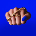 Visuel de campagne percutant pour la Maison Akillis Paris et sa collection emblématique Capture Me. Un poing fermé met en valeur un bracelet et une bague graphiques en or sertis de diamants, contrastant avec un vernis à ongles assorti au fond bleu électrique. Cliché exclusif intégré dans la section Joaillerie & Horlogerie de l'édition de Juin 2026 de THE Magazine.