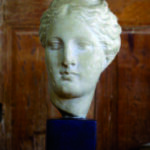 Photographie d'une pièce maîtresse de la collection Mas Ermitage - Antiques & Design à Saint-Tropez. Ce buste classique sculpté dans un marbre clair est mis en valeur sur un socle sombre devant une porte en boiserie ancienne. Cliché artistique réalisé pour l'édition de Juin 2026 de THE Magazine.