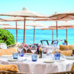 Reportage exclusif réalisé pour l'édition de Juin 2026 de THE Magazine au restaurant de plage Byblos Beach à Ramatuelle. Vue d'une élégante table ronde dressée face à la mer Méditerranée, sous les parasols emblématiques de l'établissement. Crédit photo d'origine : Alexandre Chaplier.