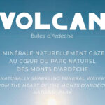 Image de couverture publicitaire pour la marque "Volcan - Bulles d'Ardèche". Le visuel présente un fond de ciel bleu azur dégradé avec un soleil lumineux en partie basse. Le texte est centré en lettres blanches, indiquant en français et en anglais : "EAU MINÉRALE NATURELLEMENT GAZEUSE AU CŒUR DU PARC NATUREL DES MONTS D’ARDÈCHE". Dans le coin inférieur droit, on aperçoit le goulot d'une bouteille en verre transparent surmontée d'une capsule bleu turquoise.