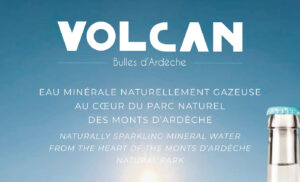 Image de couverture publicitaire pour la marque "Volcan - Bulles d'Ardèche". Le visuel présente un fond de ciel bleu azur dégradé avec un soleil lumineux en partie basse. Le texte est centré en lettres blanches, indiquant en français et en anglais : "EAU MINÉRALE NATURELLEMENT GAZEUSE AU CŒUR DU PARC NATUREL DES MONTS D’ARDÈCHE". Dans le coin inférieur droit, on aperçoit le goulot d'une bouteille en verre transparent surmontée d'une capsule bleu turquoise.