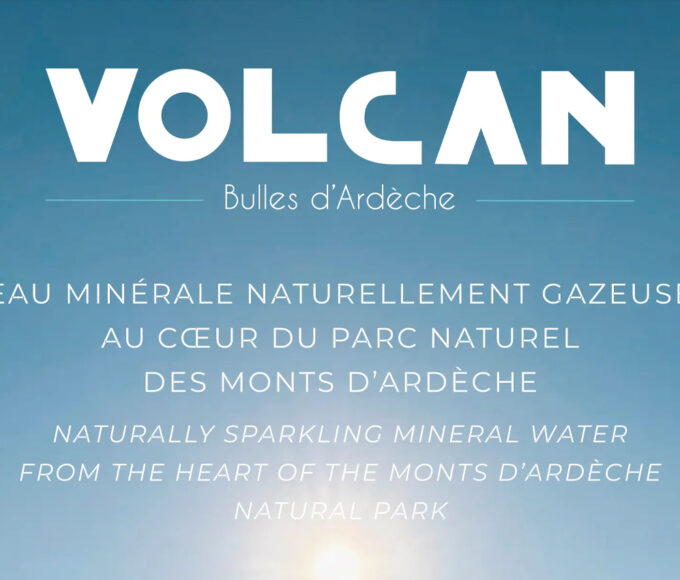 Image de couverture publicitaire pour la marque "Volcan - Bulles d'Ardèche". Le visuel présente un fond de ciel bleu azur dégradé avec un soleil lumineux en partie basse. Le texte est centré en lettres blanches, indiquant en français et en anglais : "EAU MINÉRALE NATURELLEMENT GAZEUSE AU CŒUR DU PARC NATUREL DES MONTS D’ARDÈCHE". Dans le coin inférieur droit, on aperçoit le goulot d'une bouteille en verre transparent surmontée d'une capsule bleu turquoise.