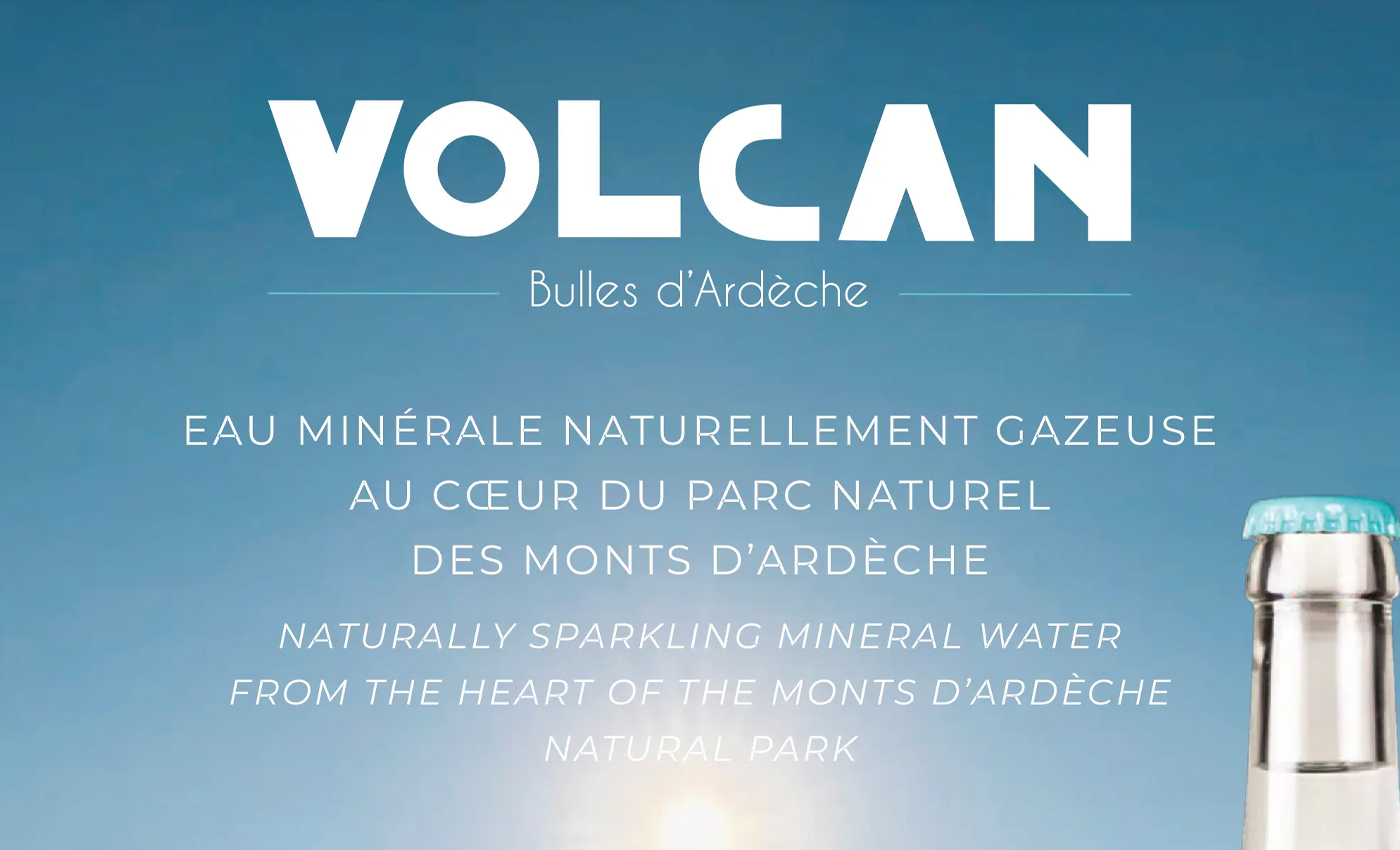 Image de couverture publicitaire pour la marque "Volcan - Bulles d'Ardèche". Le visuel présente un fond de ciel bleu azur dégradé avec un soleil lumineux en partie basse. Le texte est centré en lettres blanches, indiquant en français et en anglais : "EAU MINÉRALE NATURELLEMENT GAZEUSE AU CŒUR DU PARC NATUREL DES MONTS D’ARDÈCHE". Dans le coin inférieur droit, on aperçoit le goulot d'une bouteille en verre transparent surmontée d'une capsule bleu turquoise.