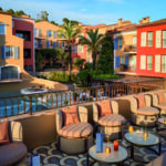 Vue plongeante depuis la terrasse supérieure de l'Hôtel Byblos à Saint-Tropez. Le premier plan met en valeur un salon d'extérieur élégant avec des fauteuils confortables aux tons saumonés posés sur un sol en mosaïque aux motifs d'arches. Des tables basses accueillent des menus bleus et des cocktails. En arrière-plan, on aperçoit la piscine de l'hôtel et les façades colorées (ocre, rouge, terre cuite) aux volets bleus qui recréent l'atmosphère unique d'un village provençal sous un ciel dégagé.