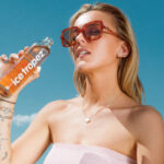 Visuel publicitaire estival pour la marque "Ice Tropez". Une jeune femme blonde portant des lunettes de soleil tendance à monture marron et un haut bustier rose clair tient une bouteille d'Ice Tropez à la main, s'apprêtant à boire. Elle arbore plusieurs bijoux fins (collier, boucles d'oreilles, bracelets) et des tatouages discrets sur l'avant-bras. L'arrière-plan présente un ciel bleu azur lumineux avec quelques nuages légers au-dessus d'une végétation méditerranéenne. Une mention légale et les coordonnées de la marque sont inscrites en blanc au bas de l'image.