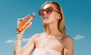 Visuel publicitaire estival pour la marque "Ice Tropez". Une jeune femme blonde portant des lunettes de soleil tendance à monture marron et un haut bustier rose clair tient une bouteille d'Ice Tropez à la main, s'apprêtant à boire. Elle arbore plusieurs bijoux fins (collier, boucles d'oreilles, bracelets) et des tatouages discrets sur l'avant-bras. L'arrière-plan présente un ciel bleu azur lumineux avec quelques nuages légers au-dessus d'une végétation méditerranéenne. Une mention légale et les coordonnées de la marque sont inscrites en blanc au bas de l'image.