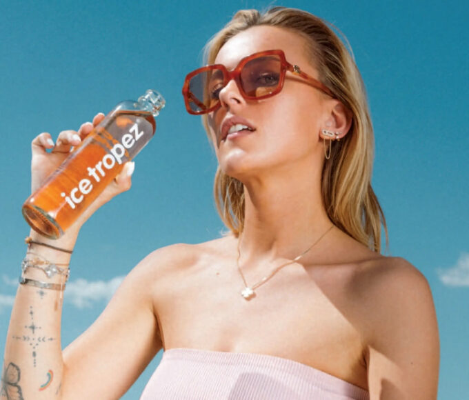 Visuel publicitaire estival pour la marque "Ice Tropez". Une jeune femme blonde portant des lunettes de soleil tendance à monture marron et un haut bustier rose clair tient une bouteille d'Ice Tropez à la main, s'apprêtant à boire. Elle arbore plusieurs bijoux fins (collier, boucles d'oreilles, bracelets) et des tatouages discrets sur l'avant-bras. L'arrière-plan présente un ciel bleu azur lumineux avec quelques nuages légers au-dessus d'une végétation méditerranéenne. Une mention légale et les coordonnées de la marque sont inscrites en blanc au bas de l'image.