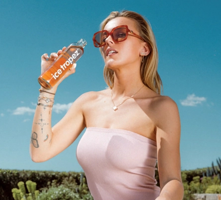 Visuel publicitaire estival pour la marque "Ice Tropez". Une jeune femme blonde portant des lunettes de soleil tendance à monture marron et un haut bustier rose clair tient une bouteille d'Ice Tropez à la main, s'apprêtant à boire. Elle arbore plusieurs bijoux fins (collier, boucles d'oreilles, bracelets) et des tatouages discrets sur l'avant-bras. L'arrière-plan présente un ciel bleu azur lumineux avec quelques nuages légers au-dessus d'une végétation méditerranéenne. Une mention légale et les coordonnées de la marque sont inscrites en blanc au bas de l'image.
