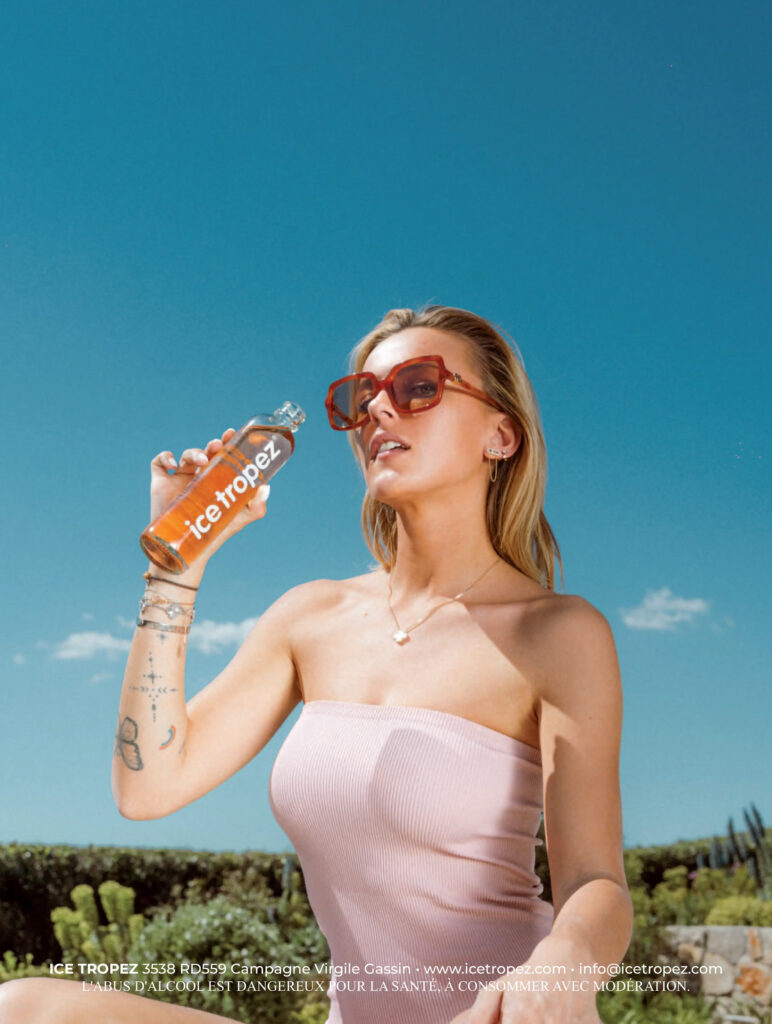 Visuel publicitaire estival pour la marque "Ice Tropez". Une jeune femme blonde portant des lunettes de soleil tendance à monture marron et un haut bustier rose clair tient une bouteille d'Ice Tropez à la main, s'apprêtant à boire. Elle arbore plusieurs bijoux fins (collier, boucles d'oreilles, bracelets) et des tatouages discrets sur l'avant-bras. L'arrière-plan présente un ciel bleu azur lumineux avec quelques nuages légers au-dessus d'une végétation méditerranéenne. Une mention légale et les coordonnées de la marque sont inscrites en blanc au bas de l'image.