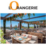 Reportage exclusif réalisé pour l'édition de Juin 2026 de THE Magazine au restaurant de plage L'Orangerie. Vue de la terrasse aménagée face à la mer avec sa pergola en bois fleurie de touches orange et ses tables dressées élégamment.
