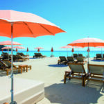 Reportage exclusif réalisé pour l'édition de Juin 2026 de THE Magazine au restaurant de plage L'Orangerie. Vue de la terrasse aménagée face à la mer avec sa pergola en bois fleurie de touches orange et ses tables dressées élégamment.