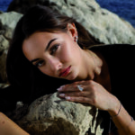 Portrait rapproché d'un jeune mannequin aux cheveux bruns posant en extérieur en bord de mer. Appuyée contre des rochers clairs, elle porte une tenue noire et met en valeur plusieurs bijoux scintillants de la Boutique Eco Diamant : une bague de style "Toi et Moi" avec deux diamants imposants (dont un en forme de cœur), des boucles d'oreilles assorties, une fine chaîne de cou et un bracelet rivière étincelant. L'arrière-plan montre la mer bleue et des formations rocheuses.