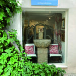 Reportage exclusif réalisé pour l'édition de Juin 2026 de THE Magazine à la boutique Mas Ermitage - Antiques & Design à Saint-Tropez. Vue de la vitrine élégante encadrée par un figuier luxuriant, mettant en scène des pièces uniques, une vasque en pierre et des fauteuils raffinés.