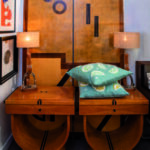 Ambiance raffinée mettant en scène du mobilier d'exception chez Mas Ermitage - Antiques & Design à Saint-Tropez. Présentation de deux consoles Art Déco aux lignes galbées en bois précieux, surmontées de lampes élégantes et de coussins turquoise aux motifs d'agrumes, devant un grand panneau en marqueterie graphique. Reportage exclusif réalisé pour l'édition de Juin 2026 de THE Magazine.