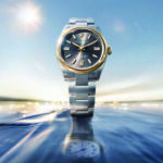 Prise de vue artistique et publicitaire d'une montre Rolex Oyster Perpetual. Le garde-temps en acier et or (Rolesor) est sublimé au lever du soleil, émergeant des flots avec son reflet parfait à la surface de l'eau. Cliché exclusif réalisé pour la section Joaillerie & Horlogerie de l'édition de Juin 2026 de THE Magazine.