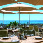 Reportage exclusif réalisé pour l'édition de Juin 2026 de THE Magazine au restaurant de plage Moorea Plage à Ramatuelle. Ambiance estivale et chic des tables dressées sur le sable, ombragées par de grands parasols face au bleu de la mer Méditerranée.