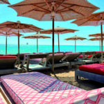 Alignement de parasols en toile tressée et de bains de soleil aux matelas graphiques sur la plage privée du Byblos Beach à Ramatuelle. Perspective estivale face à la mer turquoise pour l'édition de Juin 2026 de THE Magazine.