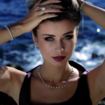 Portrait de face d'un mannequin aux cheveux relevés et yeux clairs, posant en extérieur devant un arrière-plan maritime d'un bleu profond. Elle porte une robe sombre à larges bretelles et met en valeur une parure étincelante de la Boutique Eco Diamant : un collier ras-de-cou serti de diamants de culture espacés, deux bracelets rivières de diamants (un à chaque poignet), une bague solitaire imposante à l'annulaire et des boucles d'oreilles discrètes. Ses mains sont positionnées dans ses cheveux.
