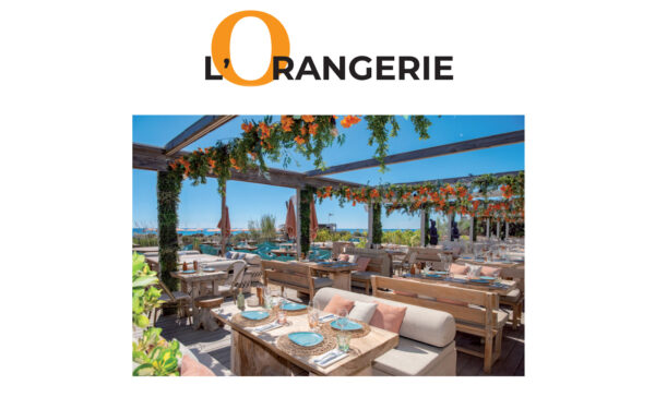 Reportage exclusif réalisé pour l'édition de Juin 2026 de THE Magazine au restaurant de plage L'Orangerie. Vue de la terrasse aménagée face à la mer avec sa pergola en bois fleurie de touches orange et ses tables dressées élégamment.