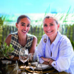 Portrait exclusif de deux femmes souriantes lors d'un déjeuner sur la terrasse du restaurant Moorea Plage à Ramatuelle. Ambiance chic, lumineuse et décontractée face à la mer. Shooting réalisé pour l'édition de Juin 2026 de THE Magazine.