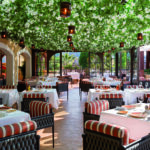 Vue sur la salle de restaurant élégamment dressée à l'Hôtel Byblos de Saint-Tropez, s'ouvrant sur une terrasse extérieure. L'espace se distingue par son plafond entièrement végétalisé et fleuri de blanc, d'où pendent des lanternes suspendues. Les tables rectangulaires portent des nappes blanches immaculées et sont entourées de fauteuils tressés aux coussins à rayures rouges et blanches. Une vaisselle colorée et des verres en cristal attendent les convives sous une lumière estivale tamisée.