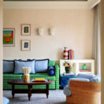 Vue sur le salon lumineux et coloré d'une suite de l'Hôtel Byblos à Saint-Tropez, encadrée par une arche au premier plan. La pièce présente un grand canapé aux motifs géométriques vert et bleu, agrémenté de coussins assortis, ainsi qu'un fauteuil en osier tressé avec un rembourrage à carreaux bleus et blancs. Au centre, une table basse ronde en bois sombre repose sur un tapis texturé bleu. Le mur de fond beige est décoré de petits cadres, d'appliques design blanches et d'une console basse blanche accueillant des livres et des vases colorés.