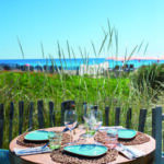 Détail d'une table ronde intimiste dressée pour le déjeuner au restaurant de plage L'Orangerie. Cadre sauvage et préservé face à la mer Méditerranée avec les dunes et barrières en bois en arrière-plan. Shooting pour l'édition de Juin 2026 de THE Magazine.