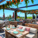 Vue panoramique exclusive de la terrasse ombragée du restaurant de plage L'Orangerie à Saint-Tropez. Tables dressées avec soin sous une pergola de bois ornée de fleurs orange, offrant une perspective sur la mer Méditerranée. Reportage réalisé pour l'édition de Juin 2026 de THE Magazine.
