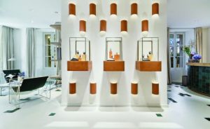 Vue intérieure exclusive de l'espace d'exposition Audemars Piguet (AP House) à Saint-Tropez. Un mur central épuré accueille trois vitrines suspendues rétroéclairées présentant les garde-temps de la manufacture, sublimées par un jeu d'appliques graphiques en terre cuite. Reportage réalisé pour la section Joaillerie & Horlogerie de l'édition de Juin 2026 de THE Magazine.