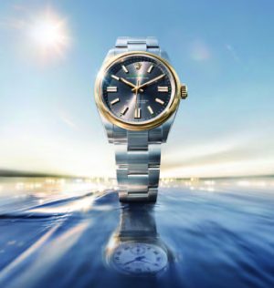 Prise de vue artistique et publicitaire d'une montre Rolex Oyster Perpetual. Le garde-temps en acier et or (Rolesor) est sublimé au lever du soleil, émergeant des flots avec son reflet parfait à la surface de l'eau. Cliché exclusif réalisé pour la section Joaillerie & Horlogerie de l'édition de Juin 2026 de THE Magazine.